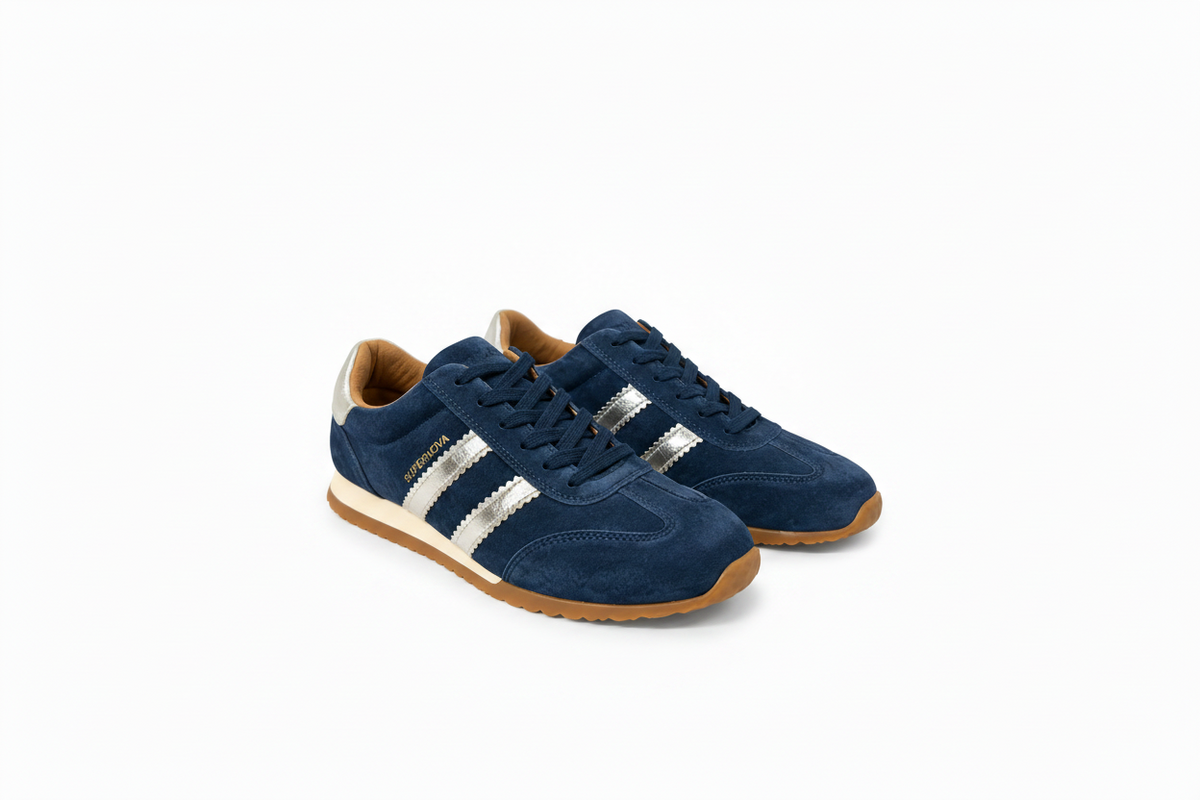 A pair of navy blue suede sneakers with metallic silver side stripes, a tan interior lining, and a brown gum sole, shown on a white background.